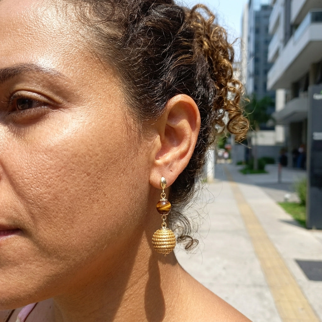 Boucles d'oreilles Jacaré en capim dourado et œil de tigre portées par une femme, vue détaillée à l'oreille en plein air