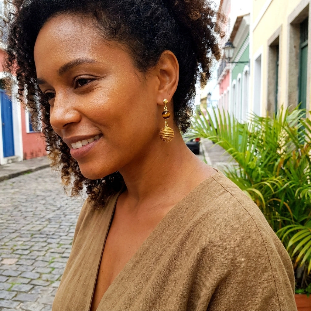 Boucles d'oreilles Jacaré en capim dourado et pierre œil de tigre portées par une femme dans une rue brésilienne, ambiance lifestyle