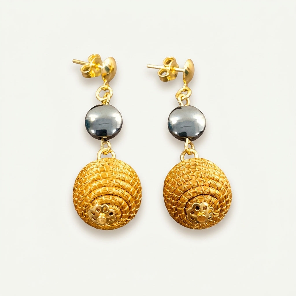 Boucles d'oreilles Taperá en capim dourado et perle hématite, sphère tressée en fibre dorée du Brésil, vue frontale sur fond clair